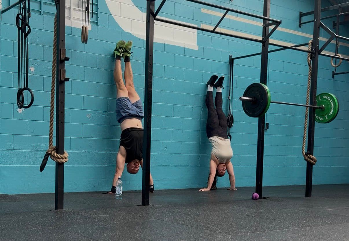 Two members practising handstands against the teal wall during open gym at CrossFit Hibiscus Coast