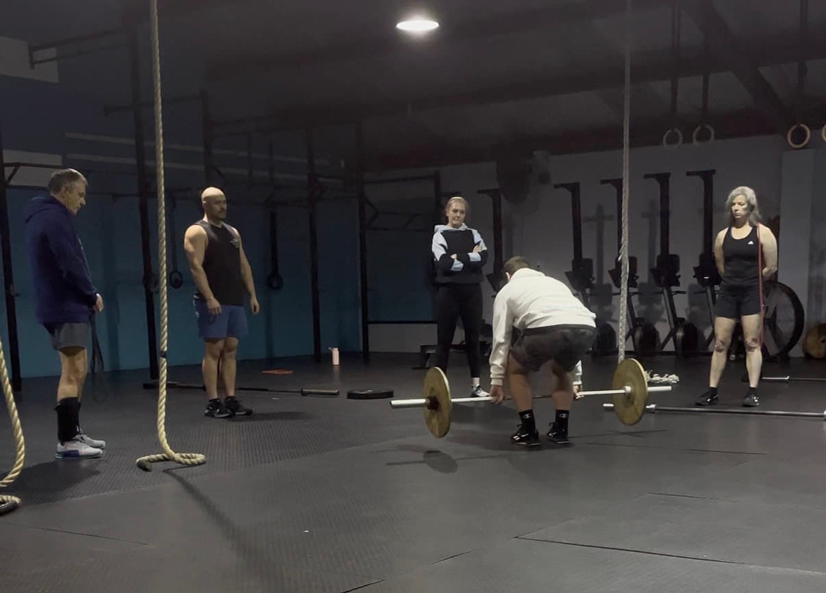 Members watching a coach demonstrate a barbell deadlift during a coached session